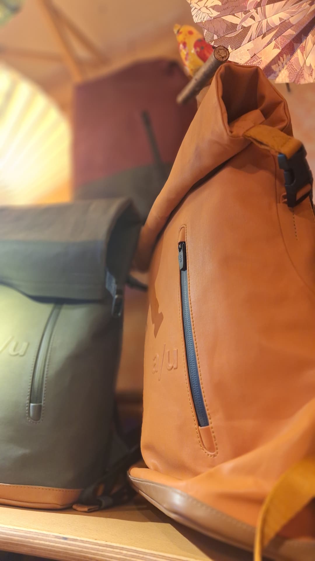 Sacs à dos a/u en cuir camel et kaki sur une étagère chez Luxiol